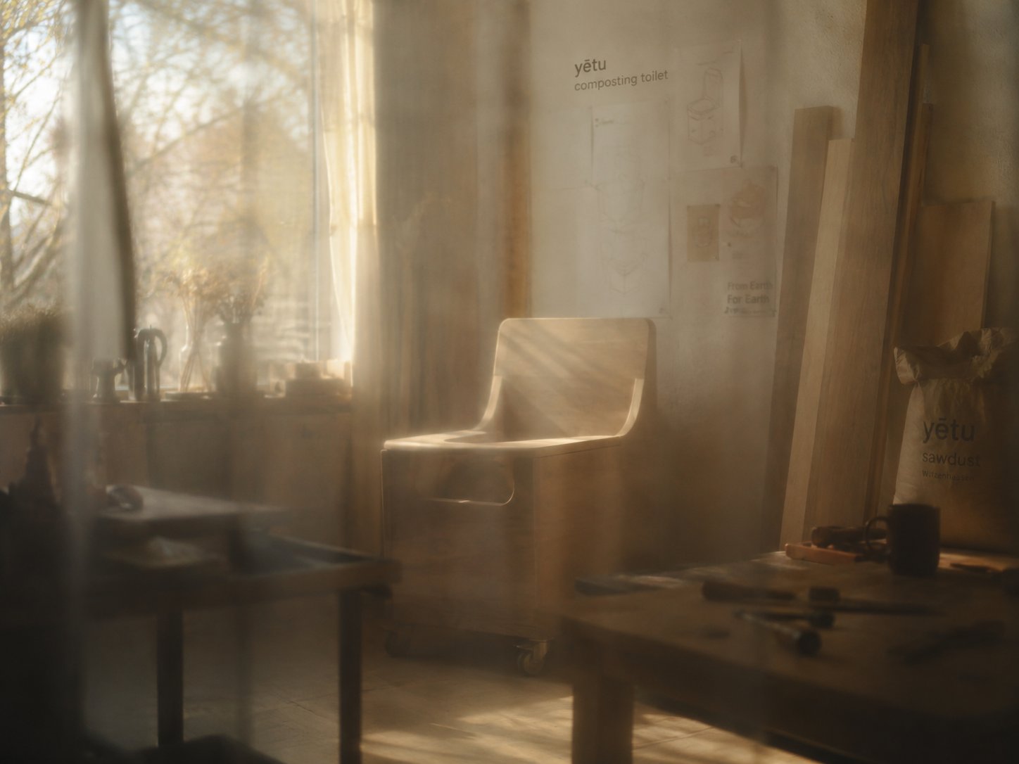 Prototype of the yētu composting toilet — a sculptural wooden seat on castors — in a sunlit workshop in Witzenhausen. Sketches and design notes on the wall, a sack of yētu sawdust beside the workbench.